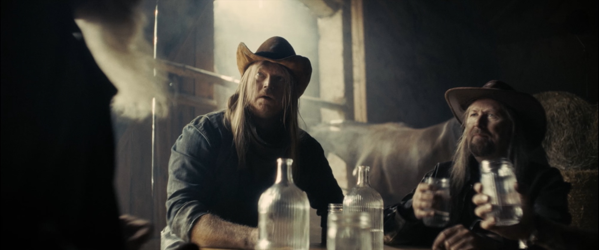 Two long-haired men in cowboy hats at a table with glass bottles inside a dusty, backlit barn - Amon Amarth